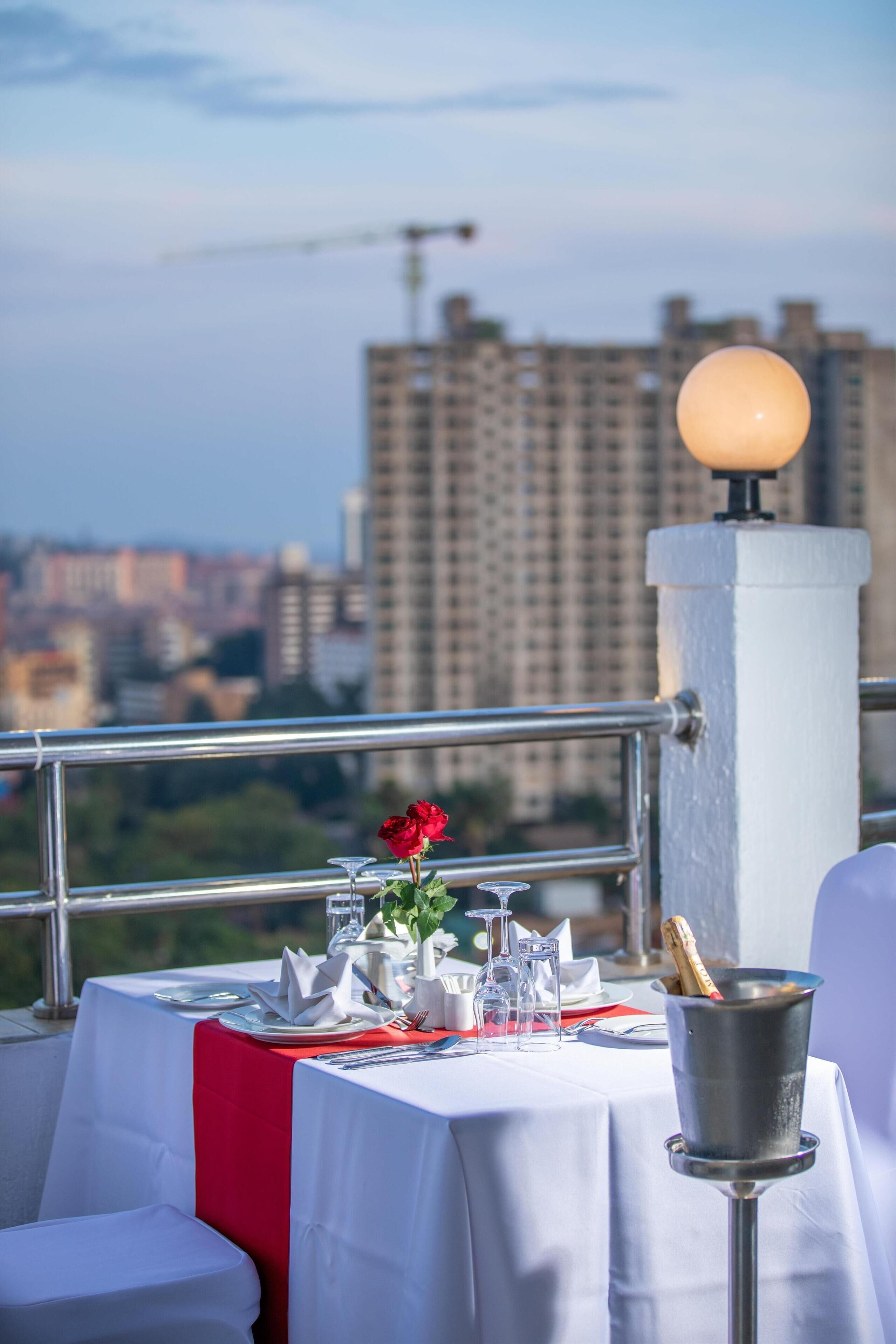 A rooftop dining area with a table set for two, a champagne bucket, and a city view in the background.