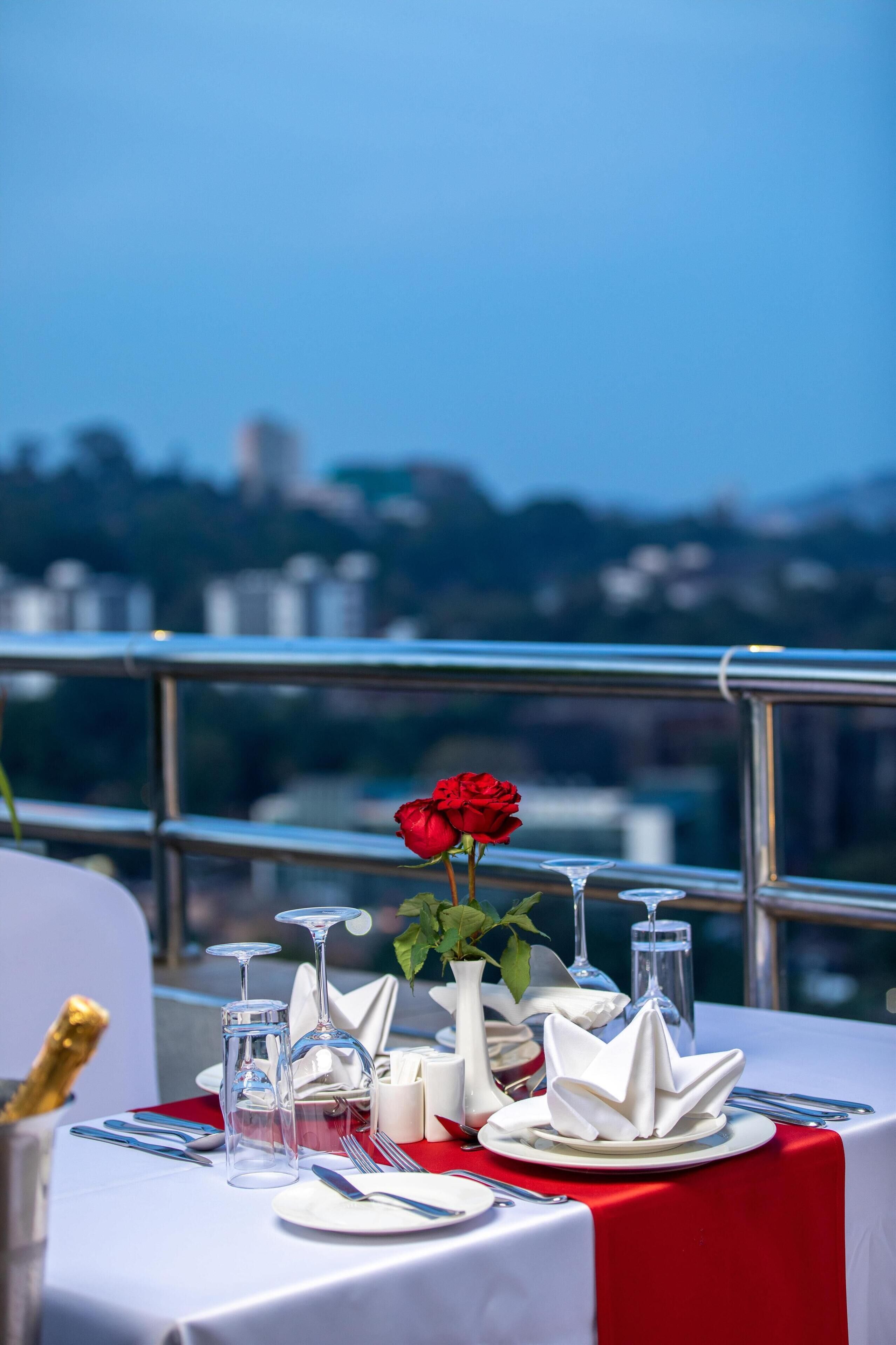 A table set for dining with a red rose centerpiece, clear glasses, and white napkins, overlooking a cityscape.