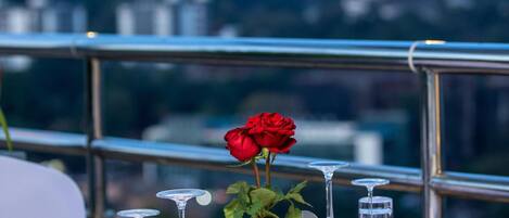 Mesa lista para comer con un centro de mesa de rosa roja, vasos transparentes y servilletas blancas, con vista a un paisaje urbano.
