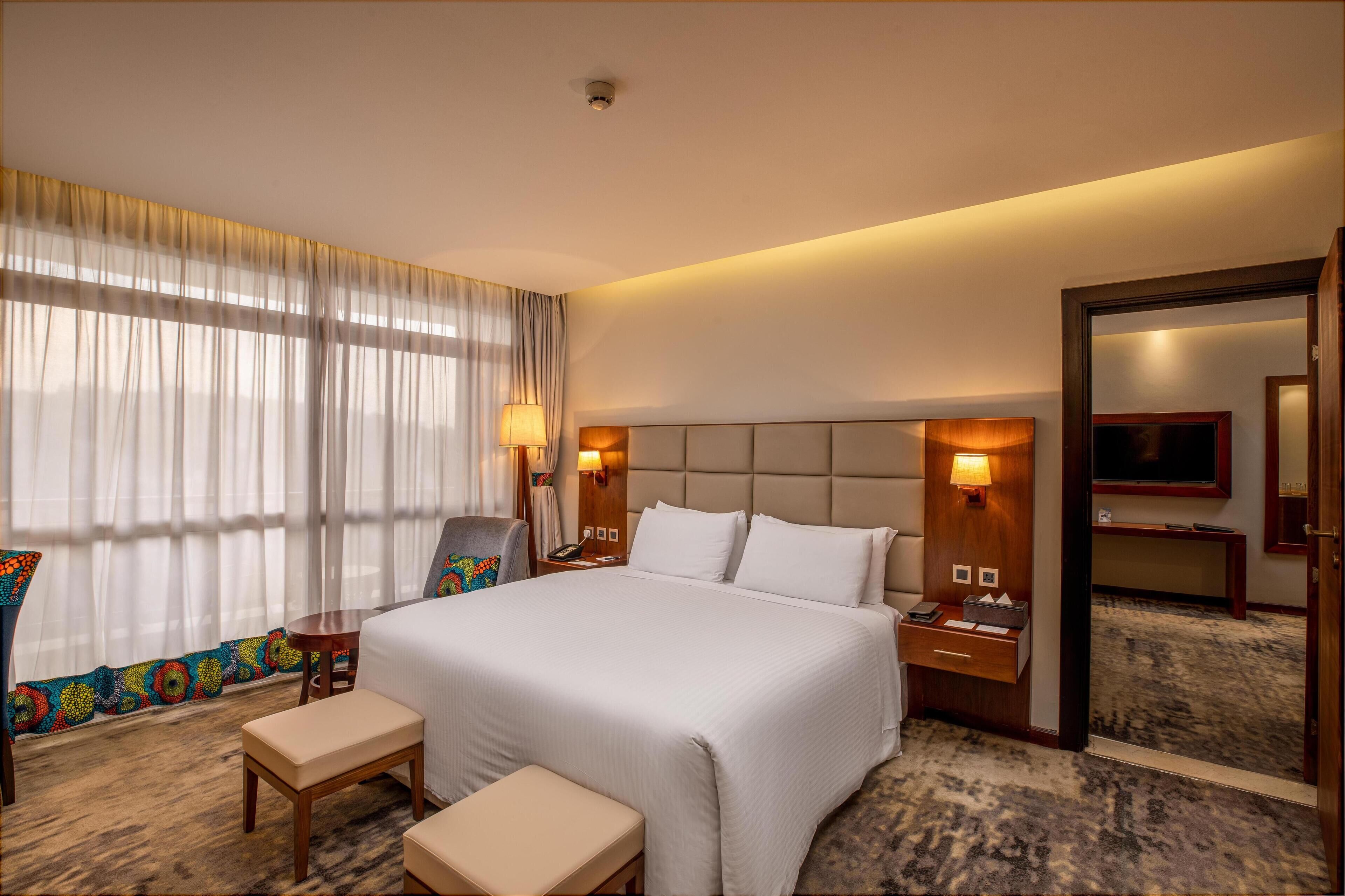 Ein Hotelzimmer mit einem großen Bett, Nachttischen, einem Sessel, einer Bank, einem Fernseher und einem Spiegel.