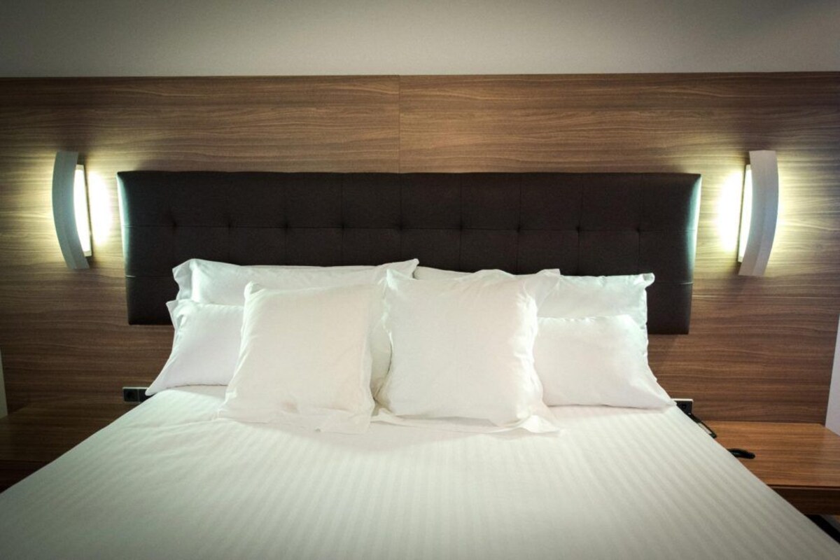 A neatly made bed with white linens and pillows, a dark headboard, and wall-mounted lighting.
