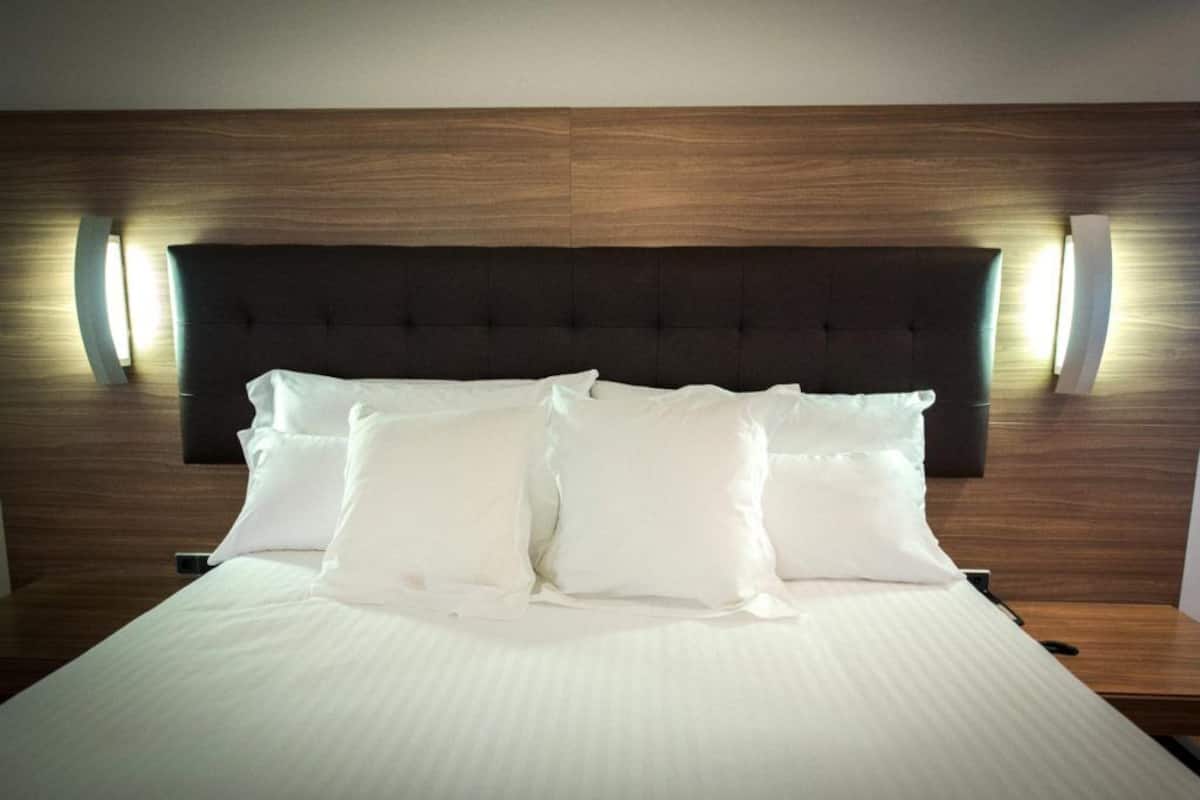 A neatly made bed with white linens and pillows, a dark headboard, and wall-mounted lighting.