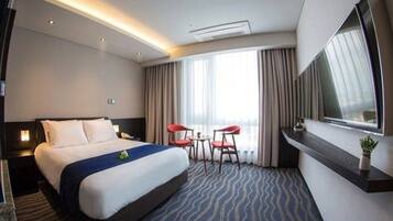A hotel room with a large bed, a desk, two chairs, a TV, and a window with curtains.