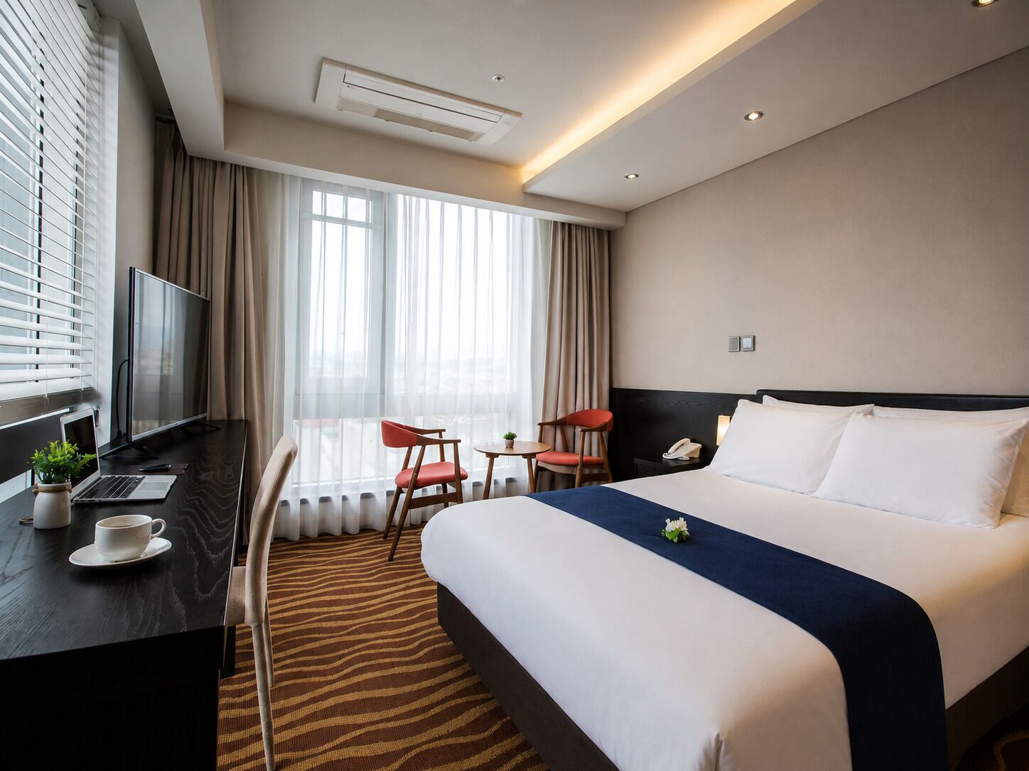 A hotel room with a bed, a desk with a laptop, a chair, a TV, and a window with curtains.