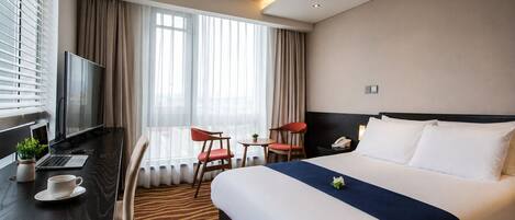 Una habitación de hotel con una cama, un escritorio con una computadora portátil, una silla, un televisor y un ventanal con cortinas.