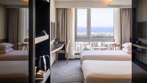 Habitación de hotel con dos camas, un escritorio, una silla, cafetera y vistas a la ciudad y al océano.