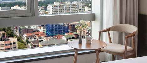 A hotel room with a large window, a bed, a chair, a small table, and a vase with flowers.