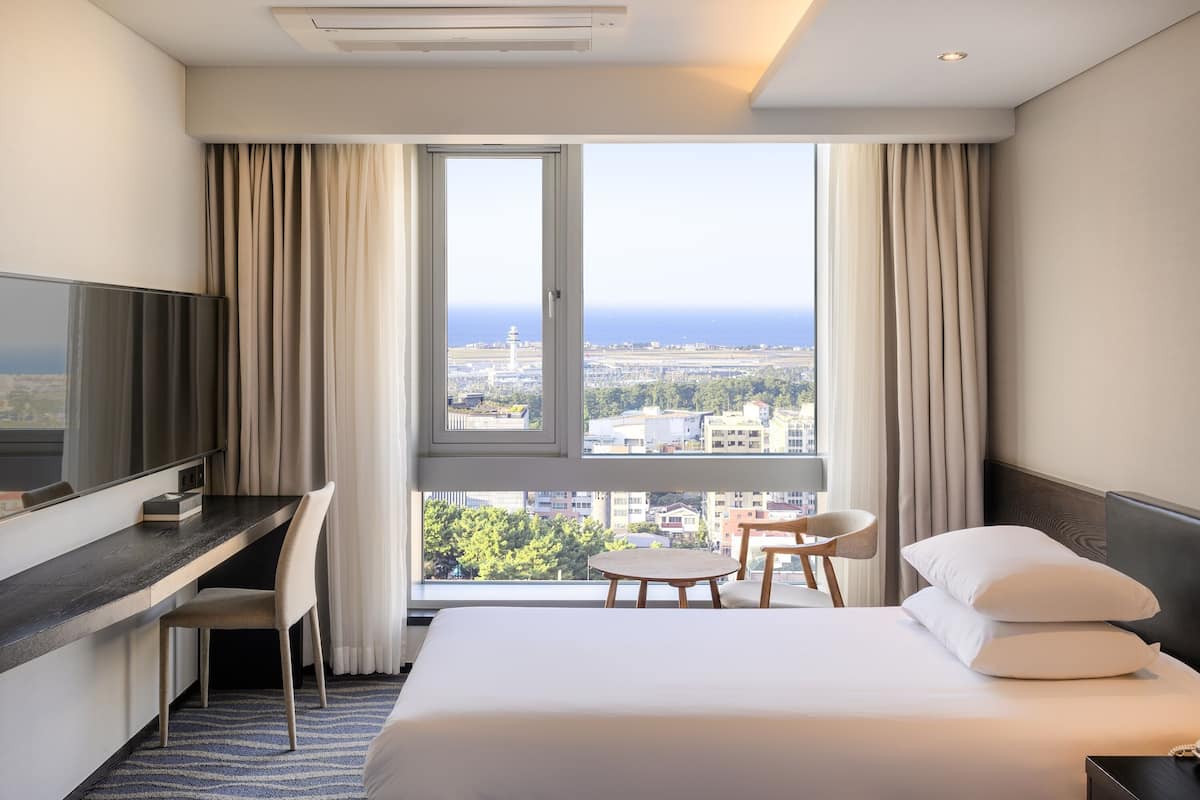 Habitación de hotel con cama, escritorio, silla, televisión y un amplio ventanal con vistas a la ciudad y al mar.