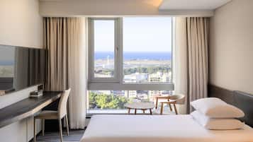 Habitación de hotel con cama, escritorio, silla, televisión y un amplio ventanal con vistas a la ciudad y al mar.