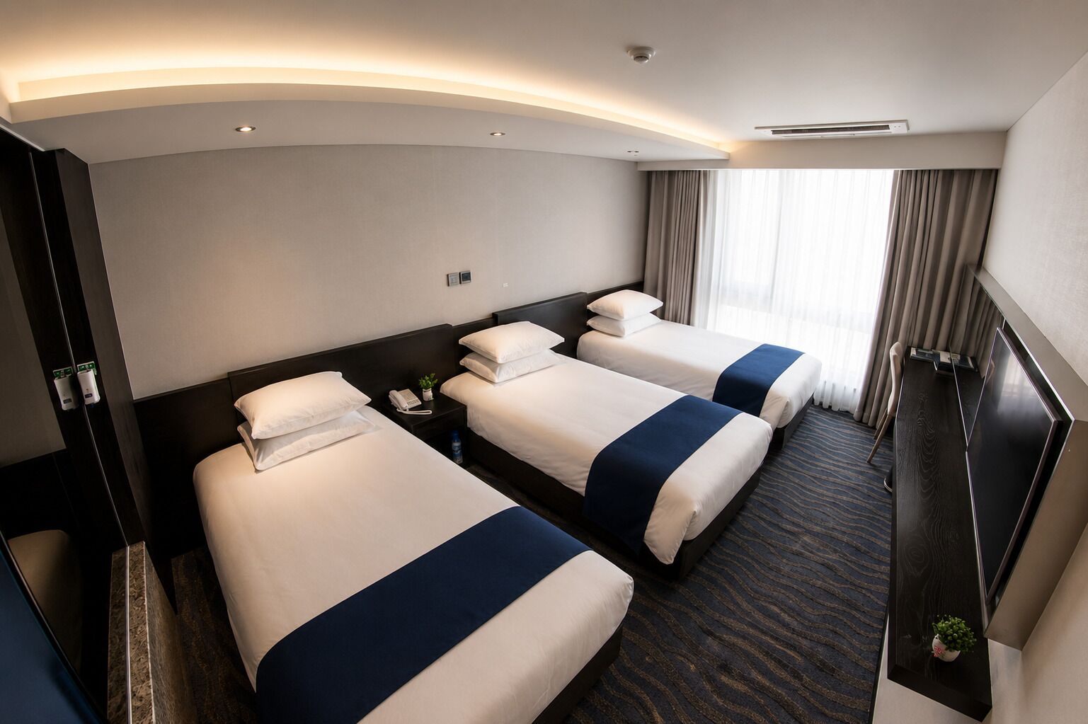 Habitación de hotel con tres camas, un escritorio, una silla y un ventanal con cortinas.