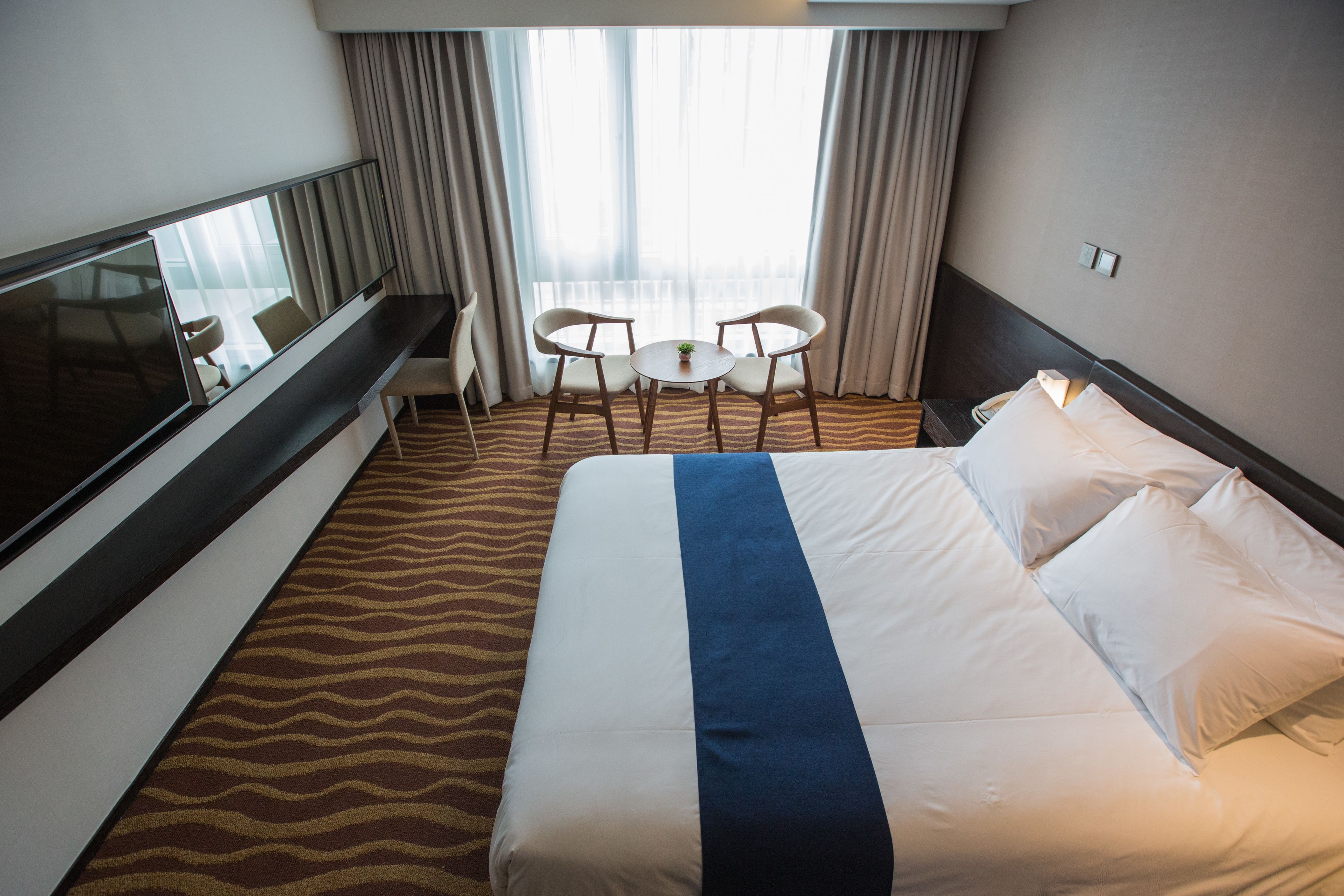 A hotel room with a large bed, a TV, a small table with two chairs, and a view of the city through the window.