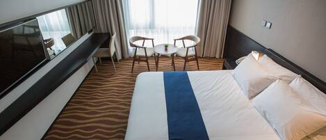 A hotel room with a large bed, a TV, a small table with two chairs, and a view of the city through the window.