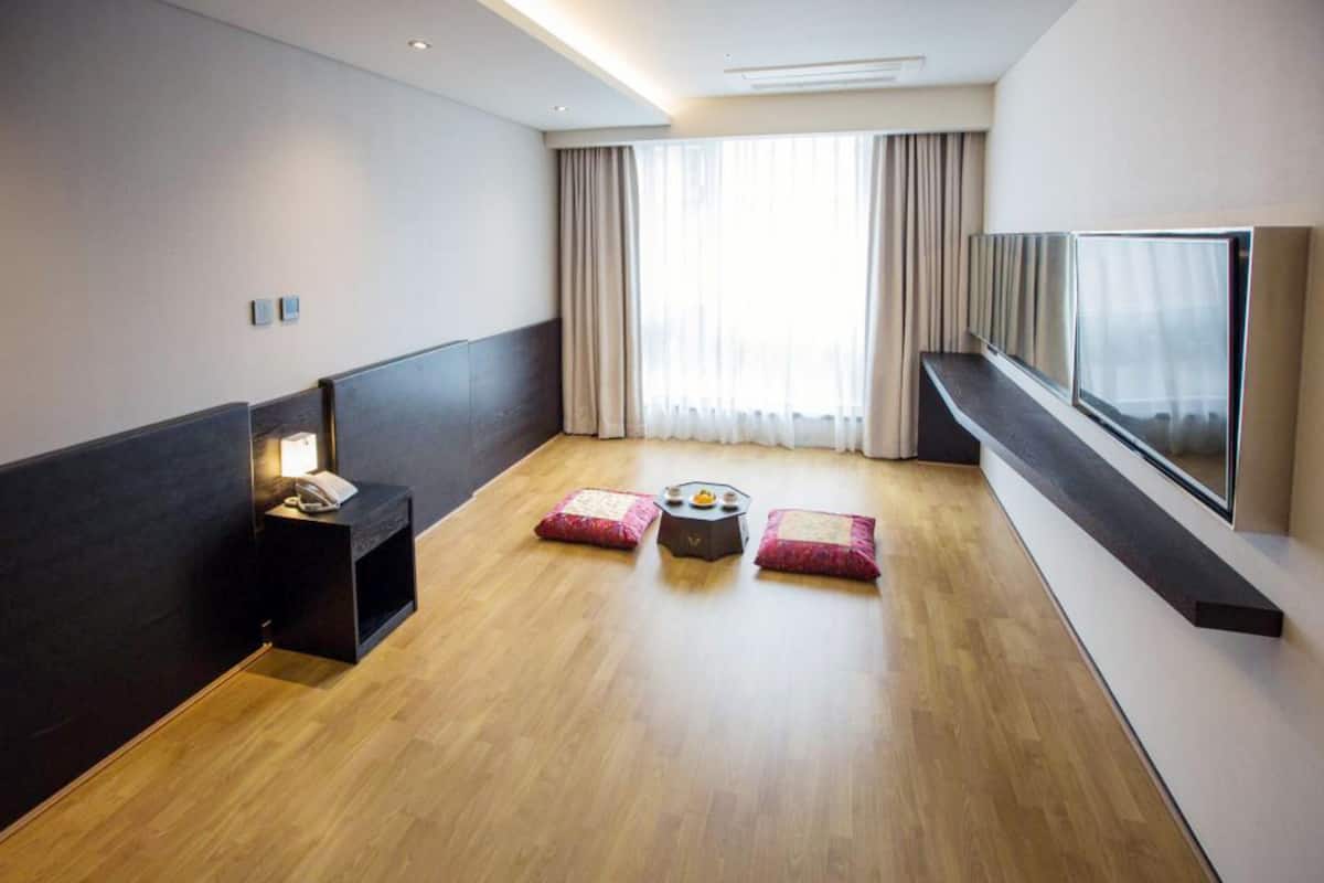 Una habitación de hotel moderna con suelo de madera, un televisor de pantalla plana montado en la pared, una mesita de noche pequeña con una lámpara y un ventanal con cortinas.