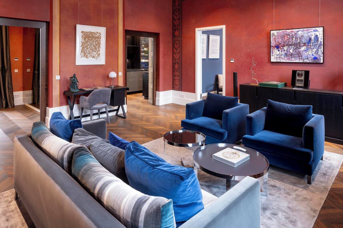 A spacious living area with a sofa, armchairs, and a coffee table. There is a kitchen in the background, a dining area, and a reception desk.