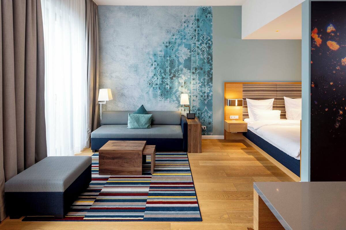 A modern hotel room with a sofa, a bed, a wooden coffee table, and a colorful striped rug.