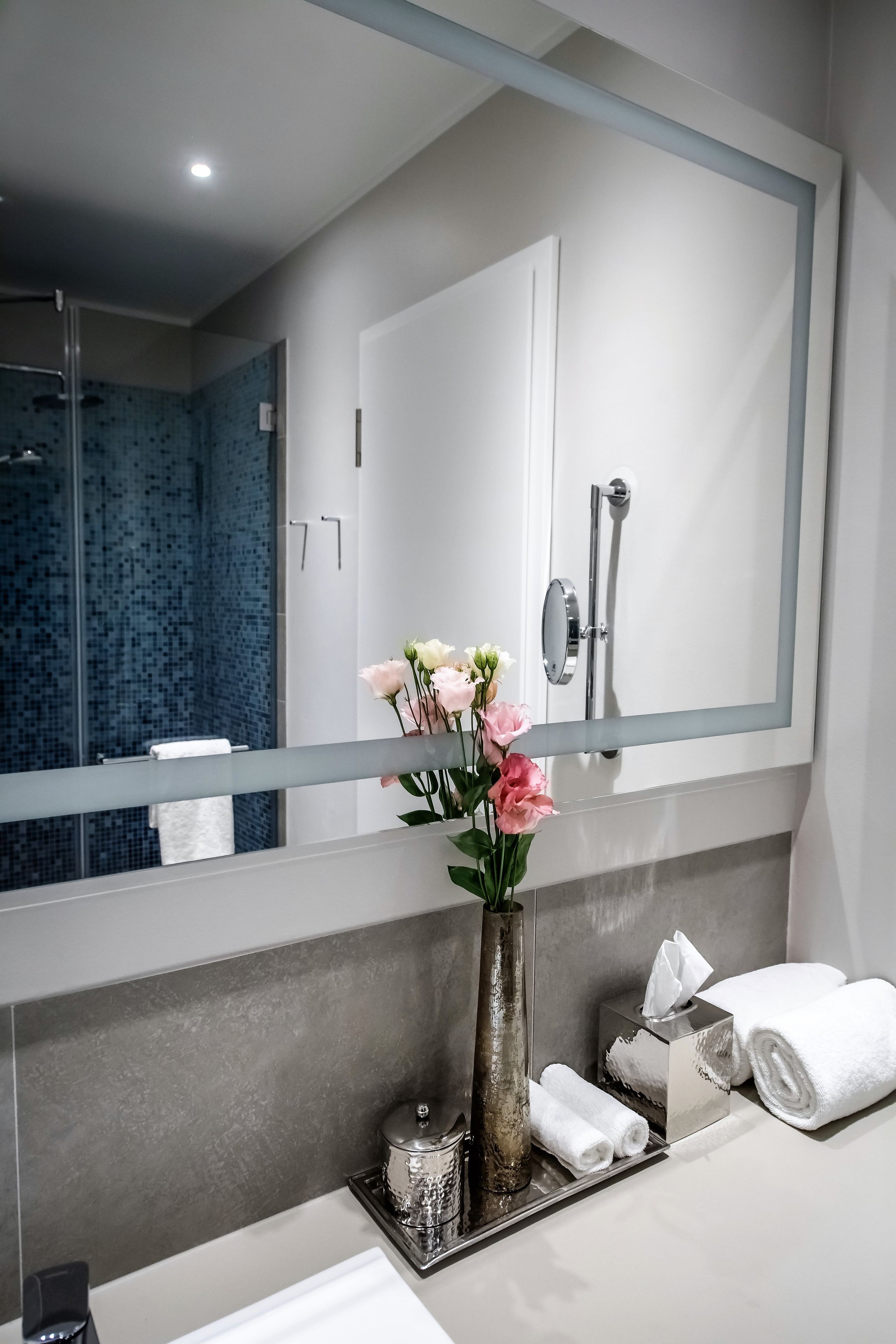 A modern bathroom with a large mirror, a shower, a vase with flowers, and neatly arranged towels.