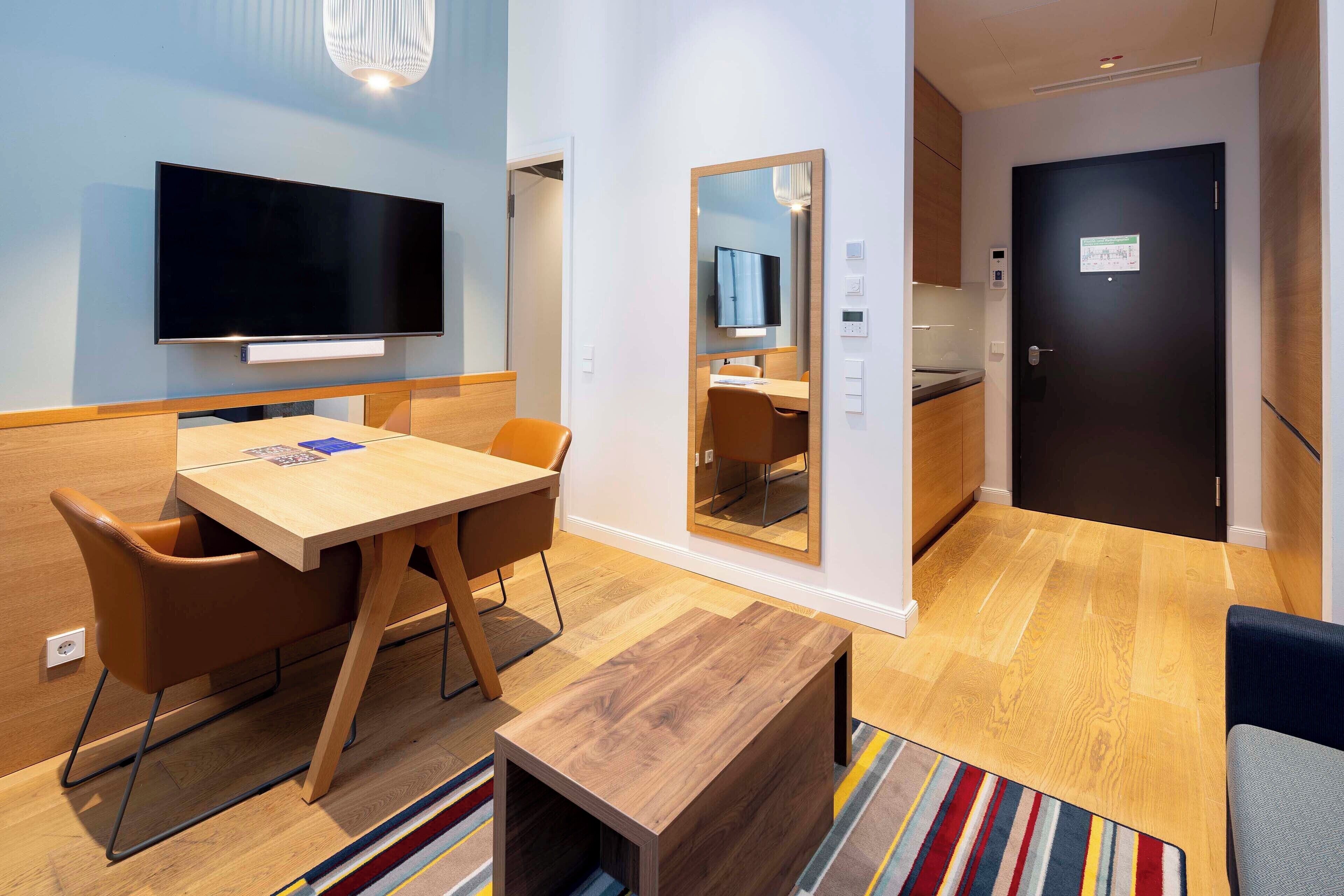 A modern hotel room with a flat-screen TV, a wooden dining table with chairs, a small wooden side table, a mirror, and a door with a sign.