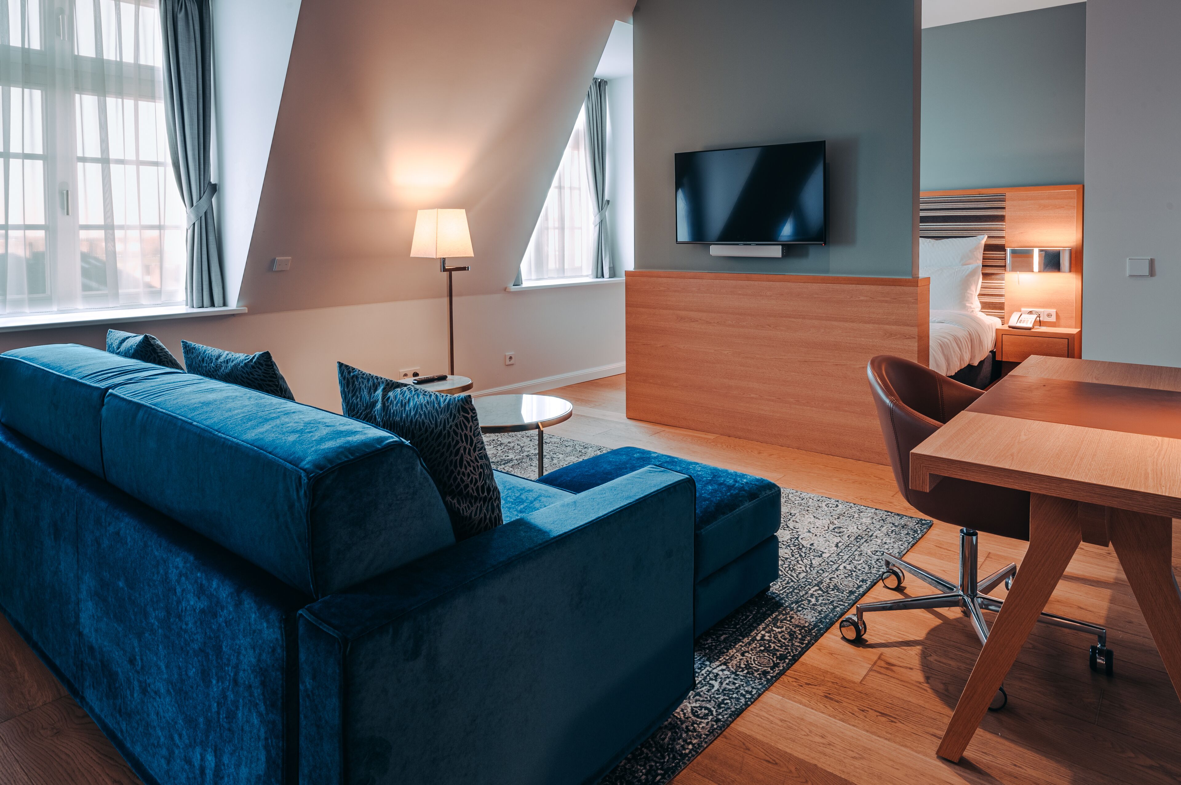 Ett modernt hotellrum med en blå soffa, ett skrivbord i trä, en platt-TV och ett litet nattduksbord med en lampa.