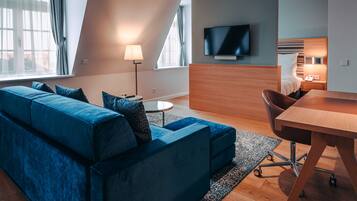 Ett modernt hotellrum med en blå soffa, ett skrivbord i trä, en platt-TV och ett litet nattduksbord med en lampa.