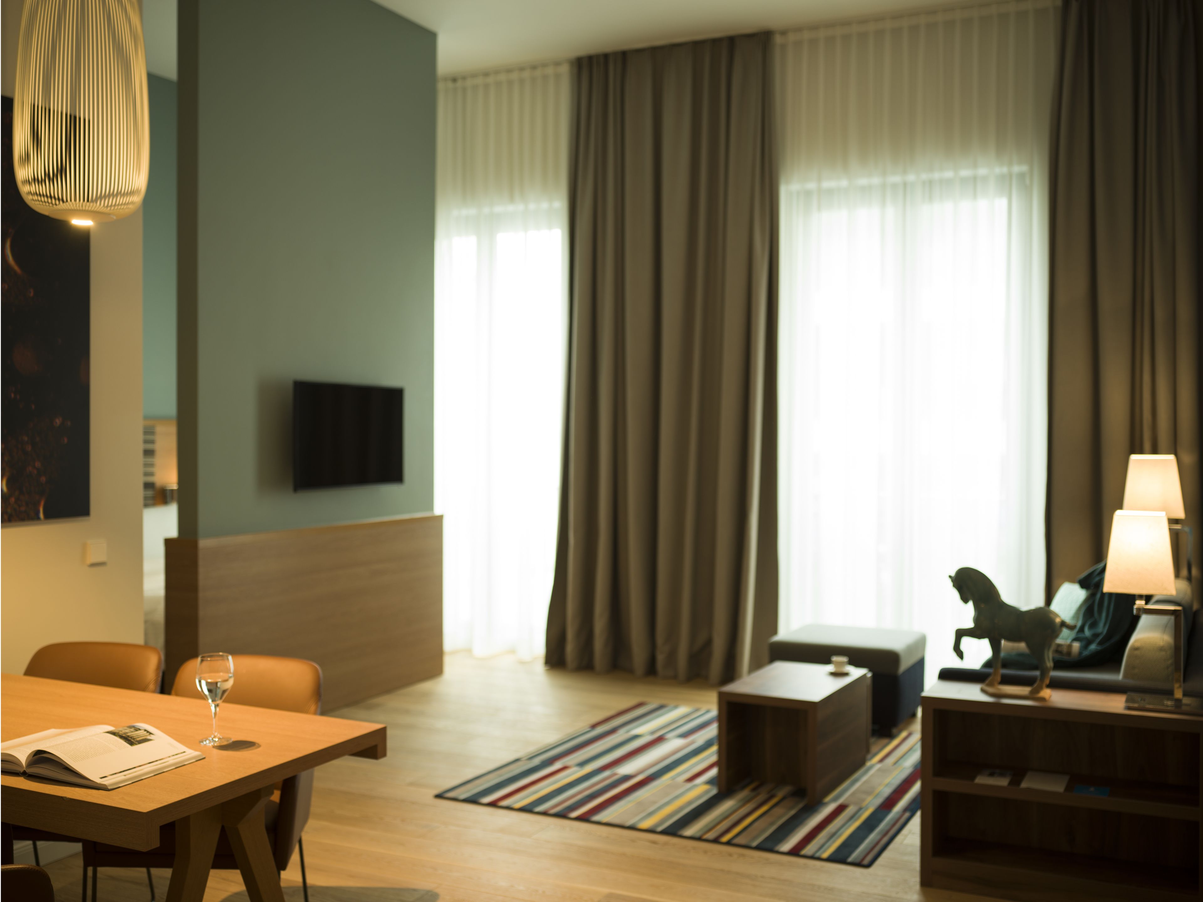 Quarto de hotel moderno com piso de madeira, tapete listrado, sofá, mesa de centro, TV montada na parede e abajur em uma mesinha lateral.