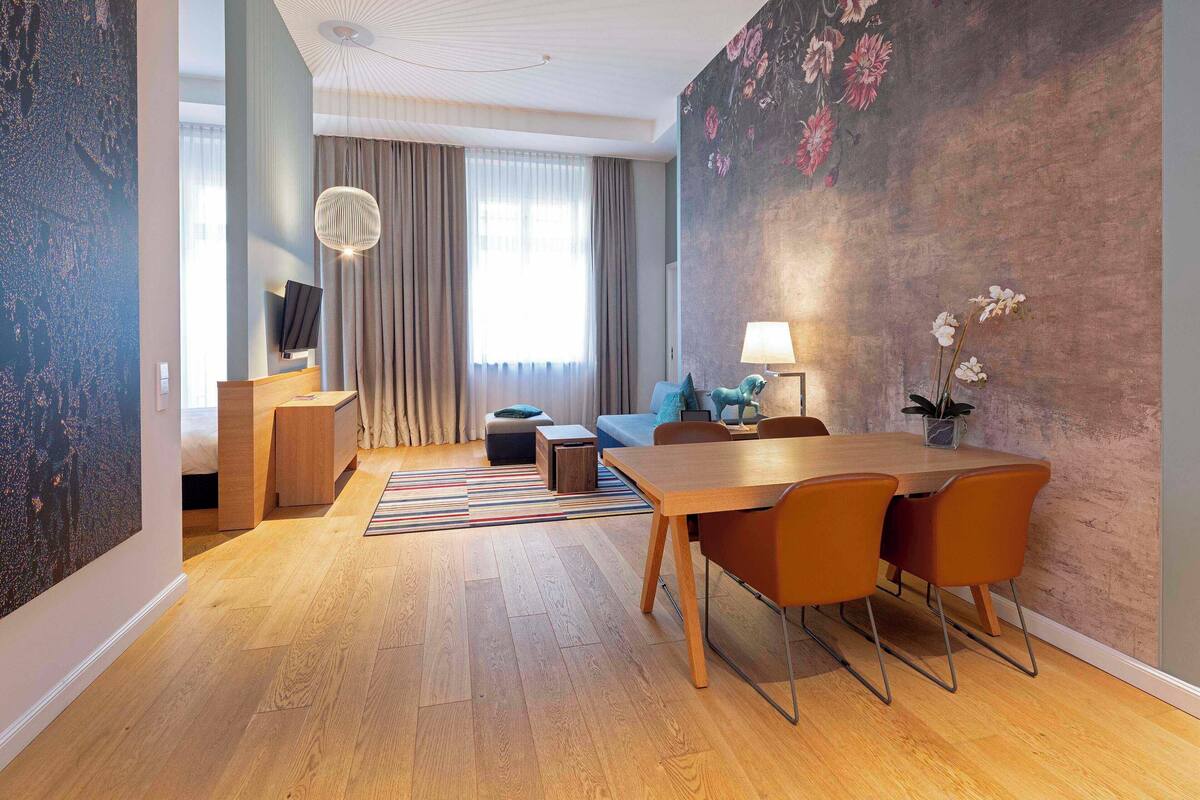 A modern living room with a wooden dining table, orange chairs, a blue sofa, and a TV mounted on the wall.