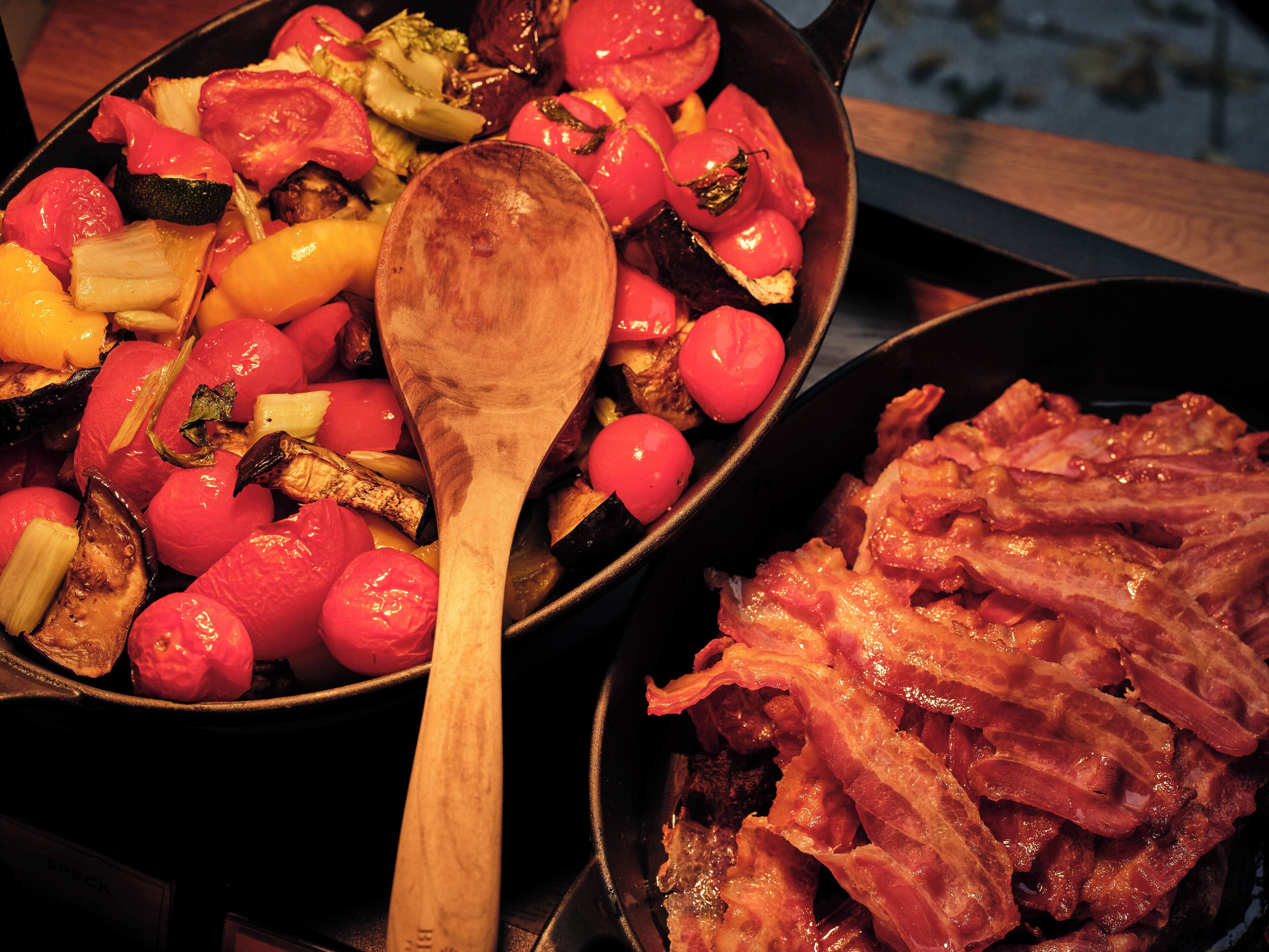Two pans of food, one with vegetables and the other with bacon, on a stove.