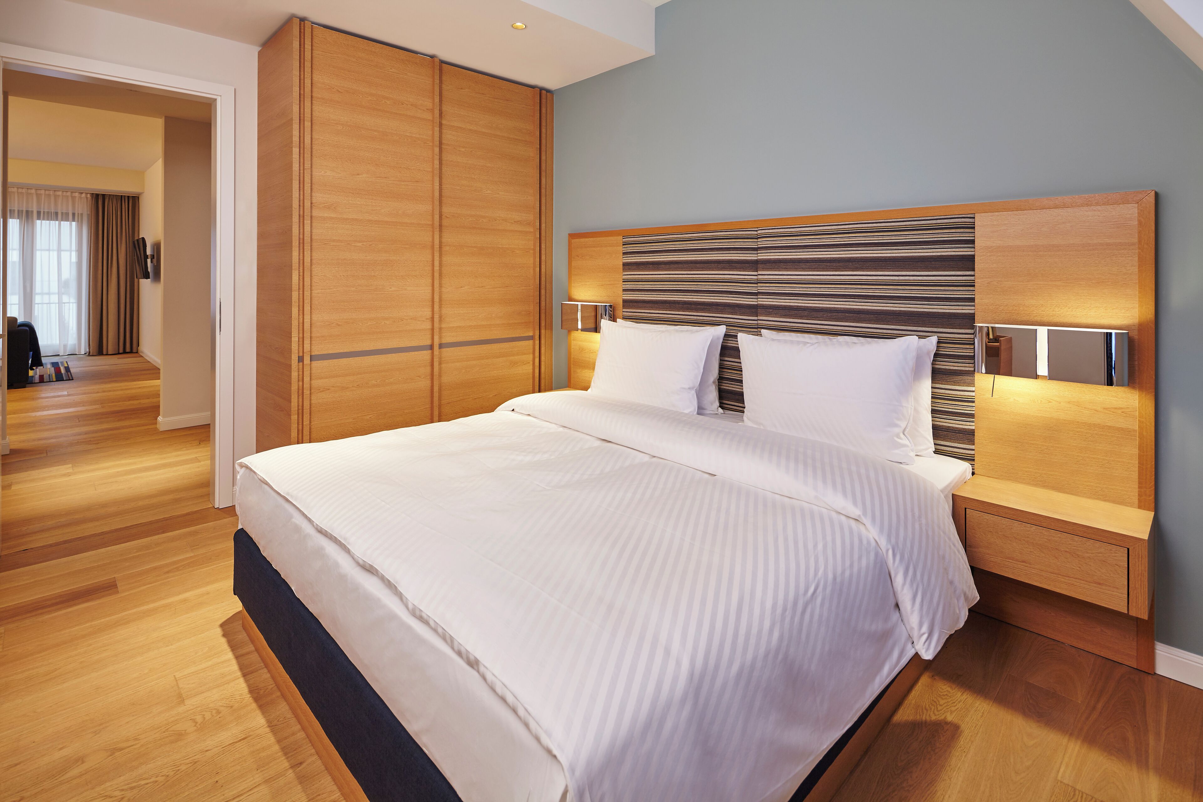 A modern hotel room with a large bed, wooden wardrobe, and a nightstand with a lamp.