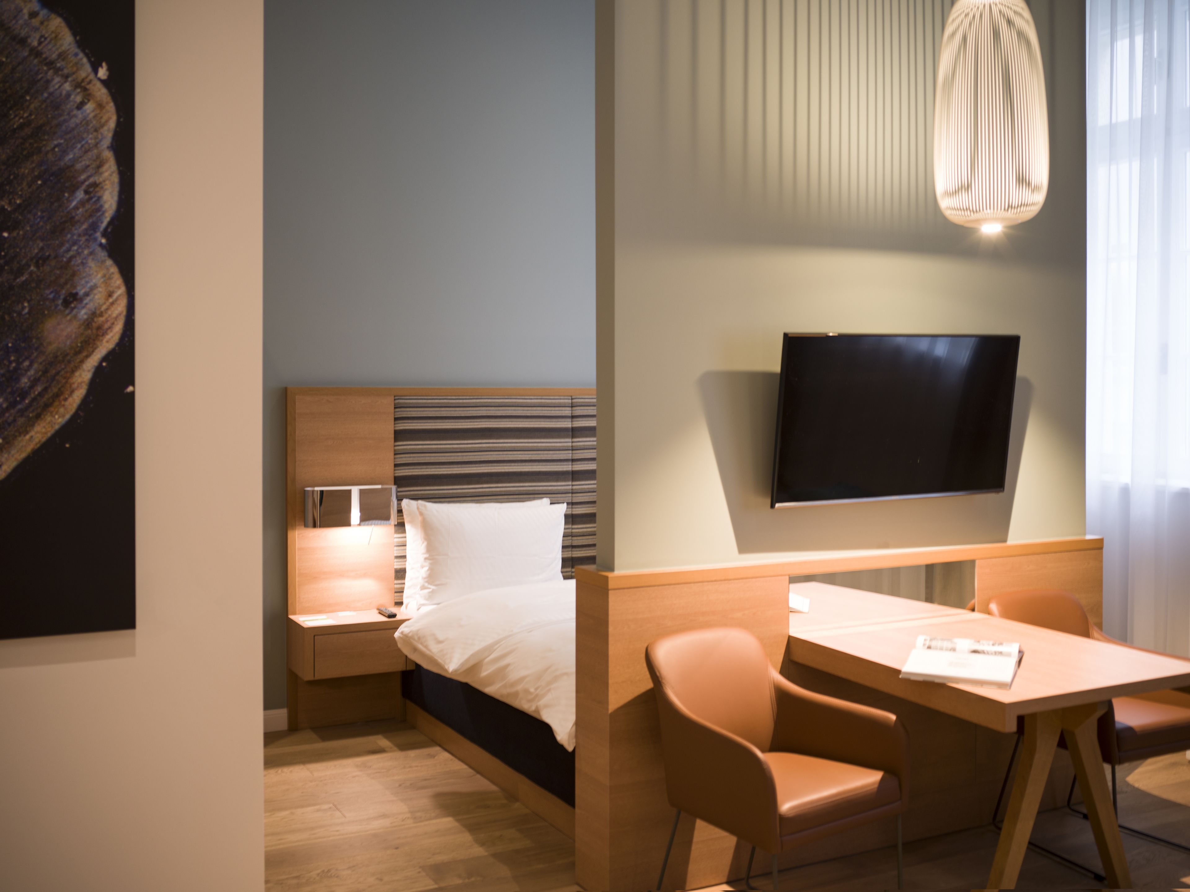 Ein modernes Hotelzimmer mit Bett, Nachttisch, Lampe, Fernseher und Schreibtisch mit Stühlen.