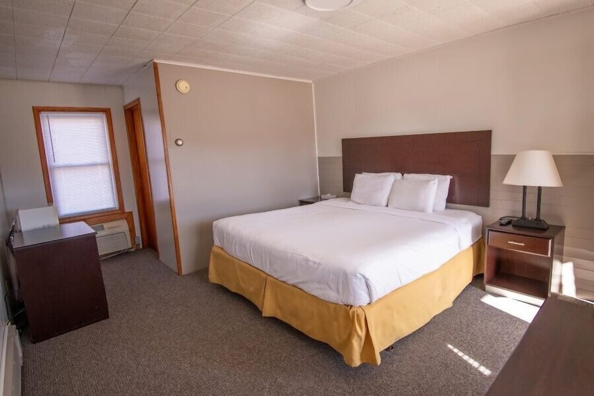 A hotel room with a large bed, a bedside table with a lamp, a desk, and a window with blinds.