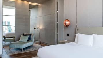 Una habitación de hotel moderna con una cama grande, un área de estar con sofá y sillones, un espejo amplio y vistas a la ciudad.