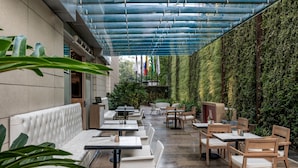 Un espace extérieur aménagé avec des bancs blancs dotés de coussins et des tables en bois, entouré d’une végétation luxuriante et surmonté d’un toit en verre.