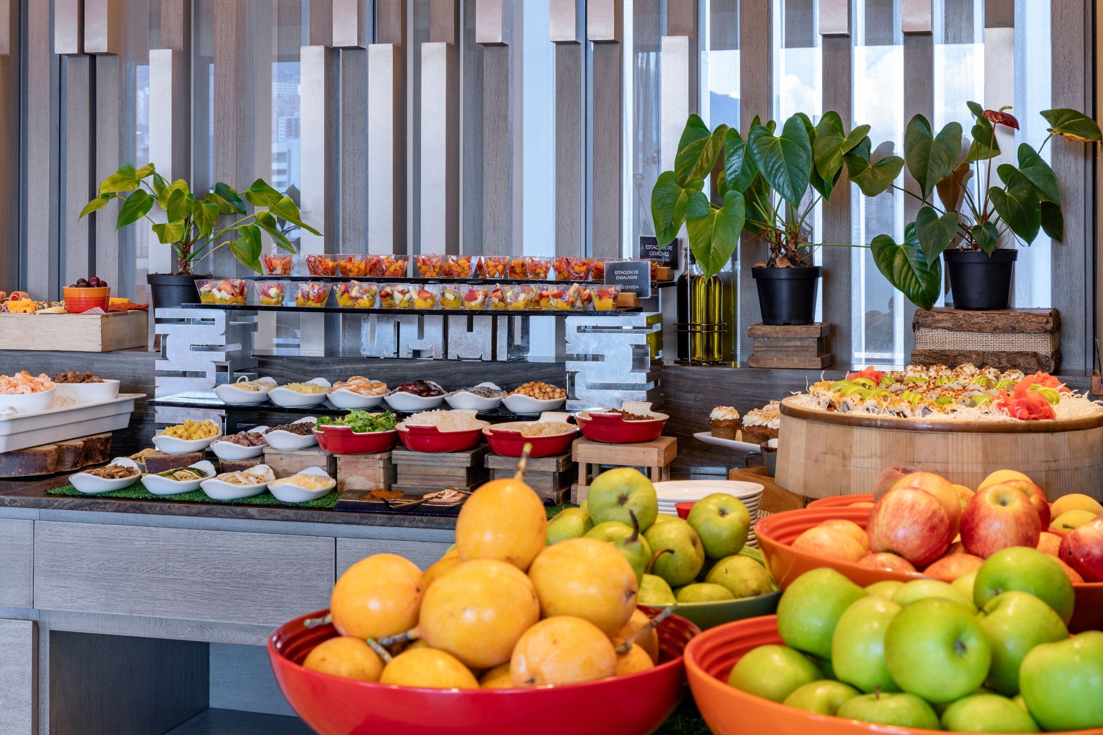 A buffet with various food items and fruits, including apples and pears, arranged on a counter with potted plants.