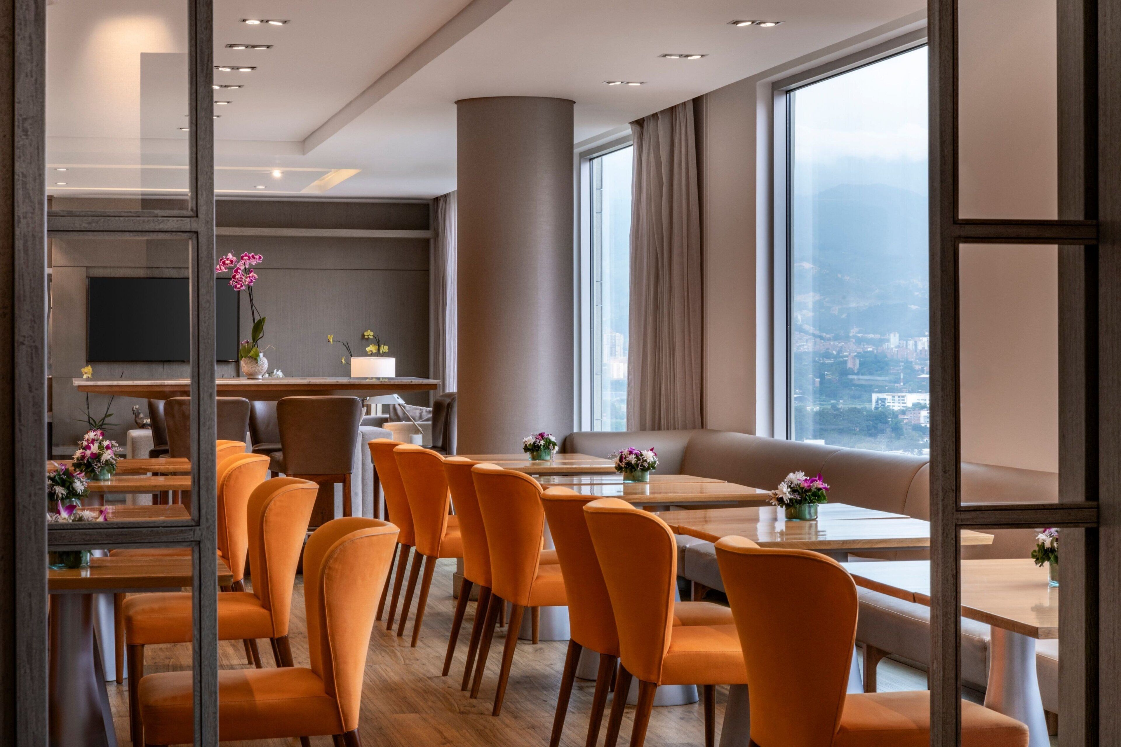 A modern dining area with orange chairs, a wooden table, and a city view through large windows.