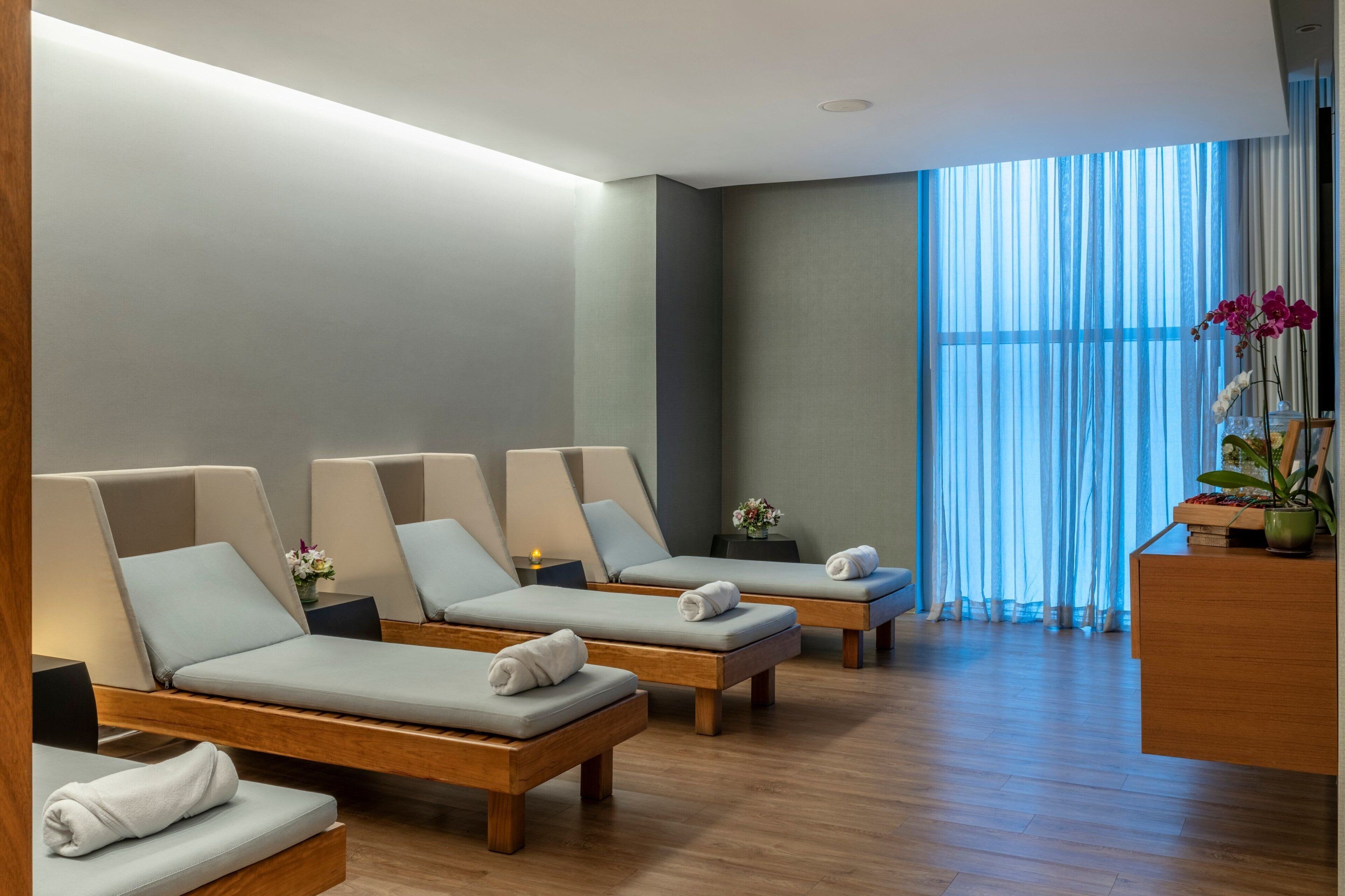 A spa room with four lounge chairs, a wooden reception desk, and a large window with sheer curtains.