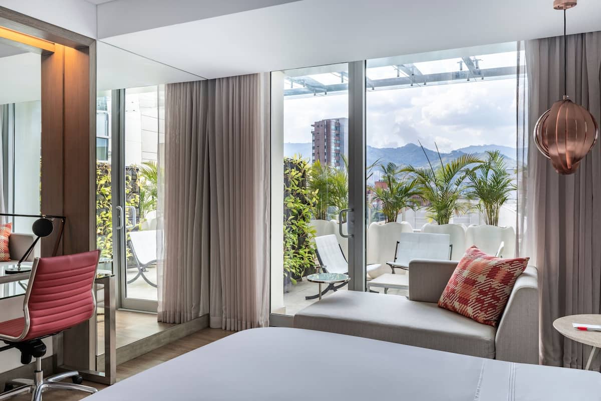 Une chambre d’hôtel moderne, dotée d’un grand lit, d’un bureau, d’une chaise et offrant une vue sur l’extérieur grâce à de grandes fenêtres.