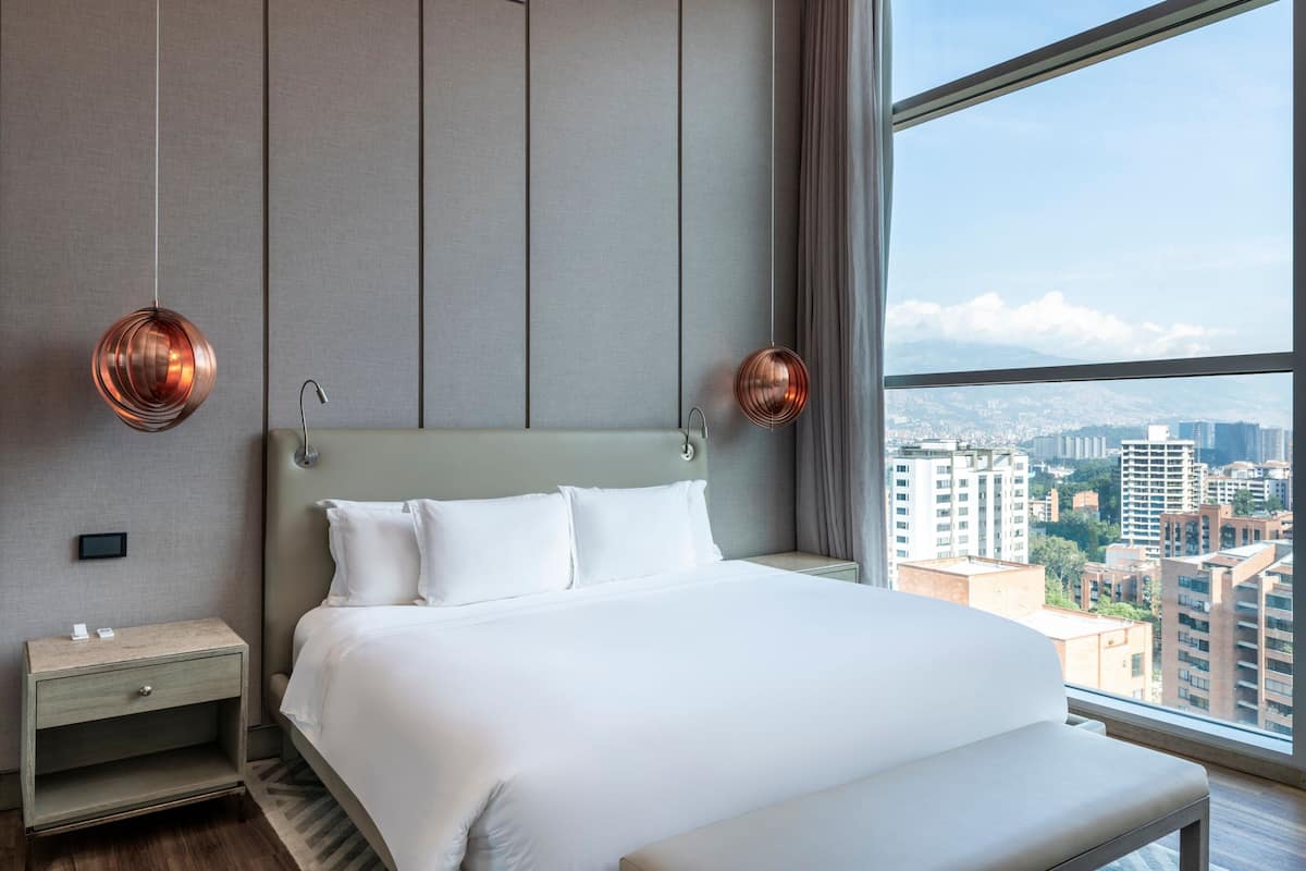 Une chambre d’hôtel moderne, dotée d’un grand lit, d’une table de chevet et offrant une vue sur la ville grâce à de grandes fenêtres.