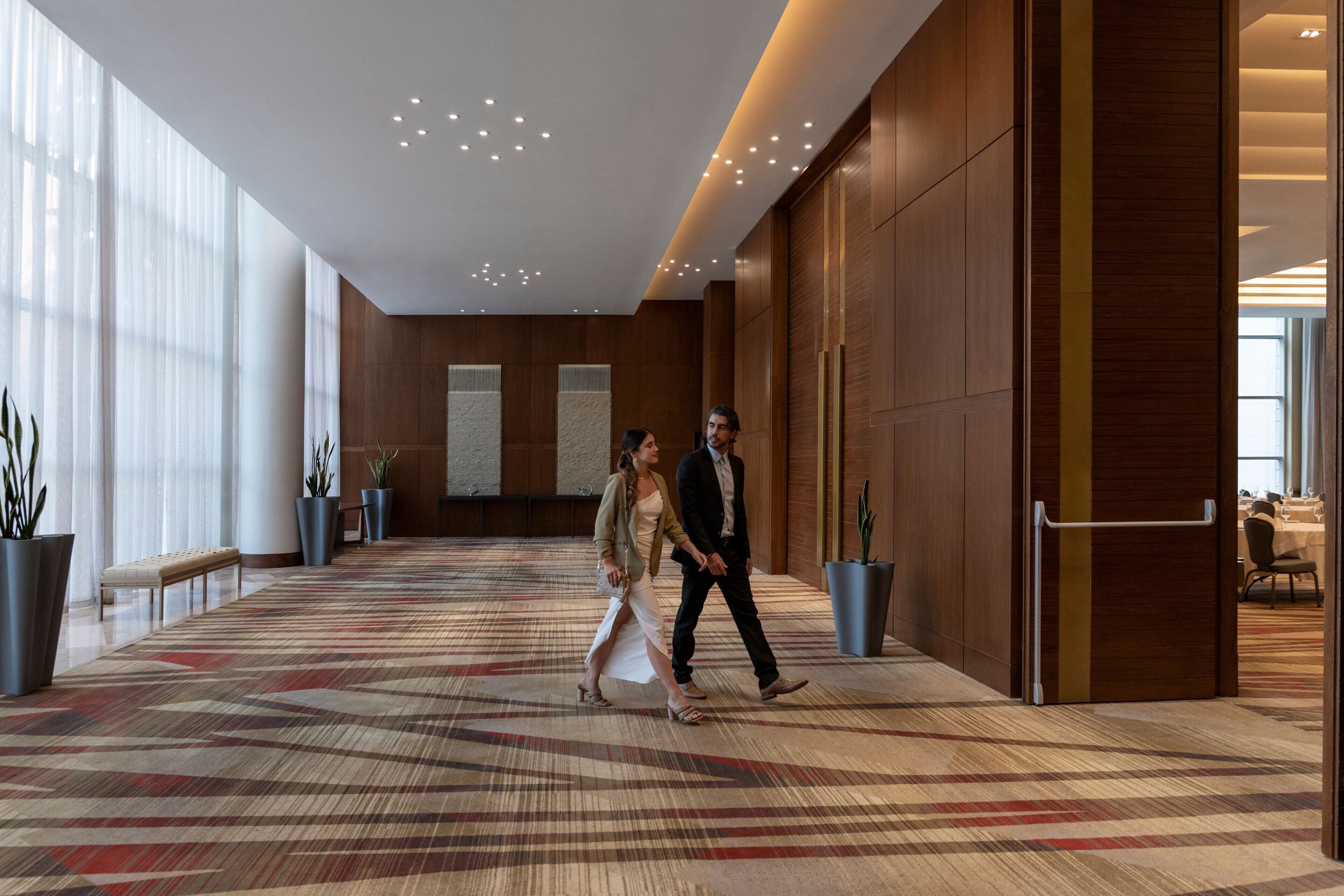 A spacious hallway with a patterned carpet, large windows, and potted plants.