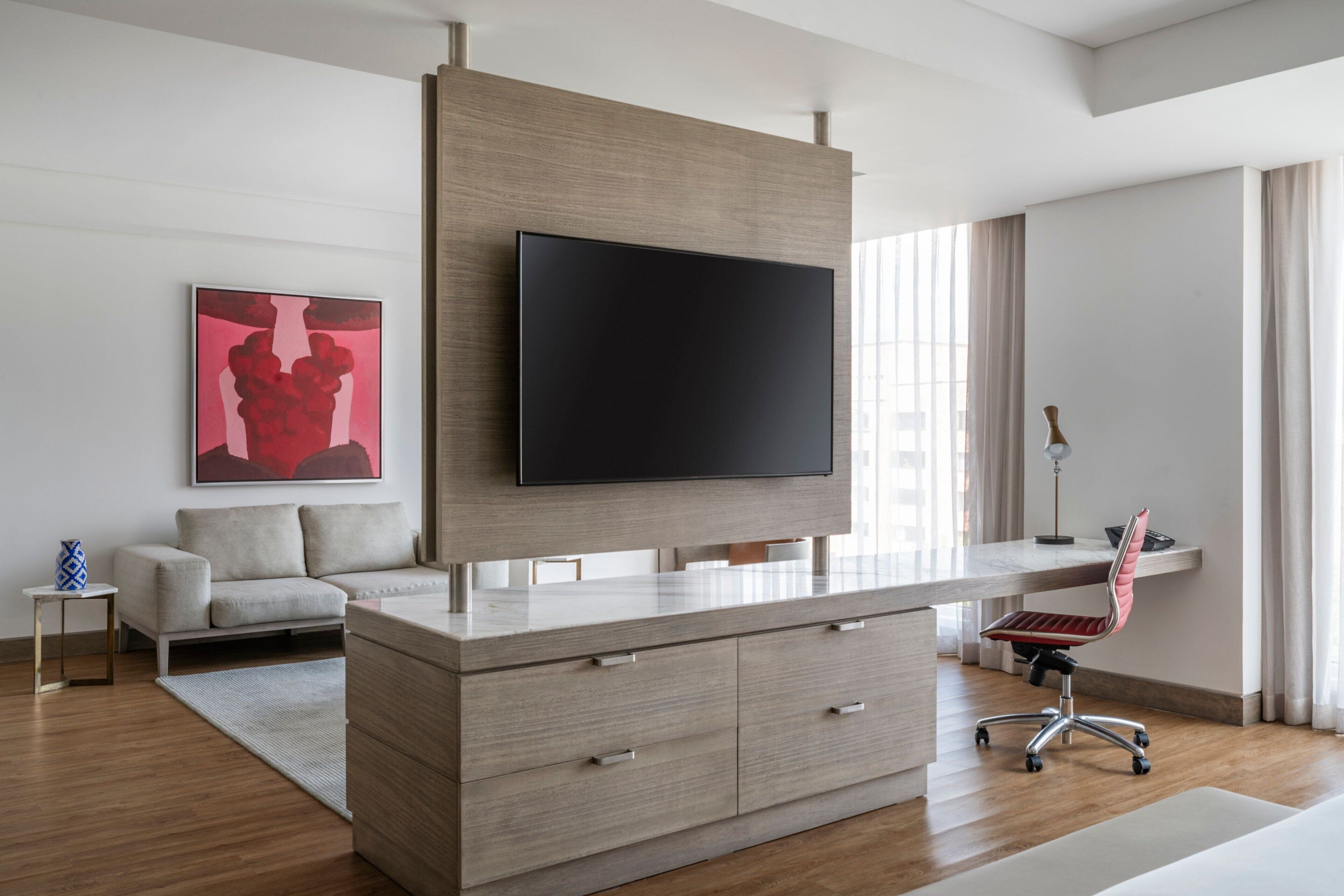 A modern living room with a large flat-screen TV mounted on a wooden wall, a grey sofa, a small side table with a vase, and a red office chair.