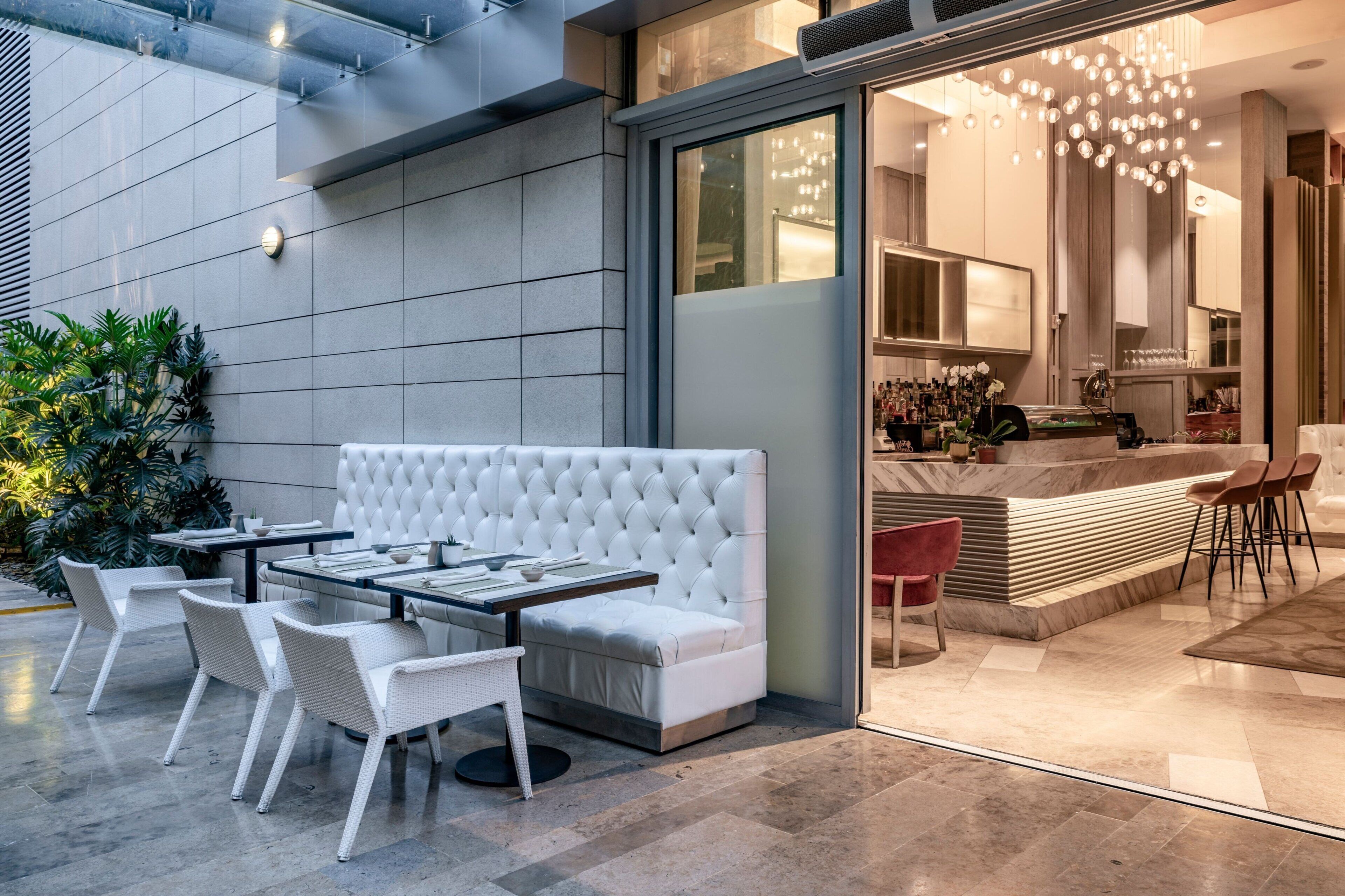 Un área de estar exterior moderna con un banco acolchado blanco y sillas de mimbre, un panel de vidrio y una zona de bar con taburetes.