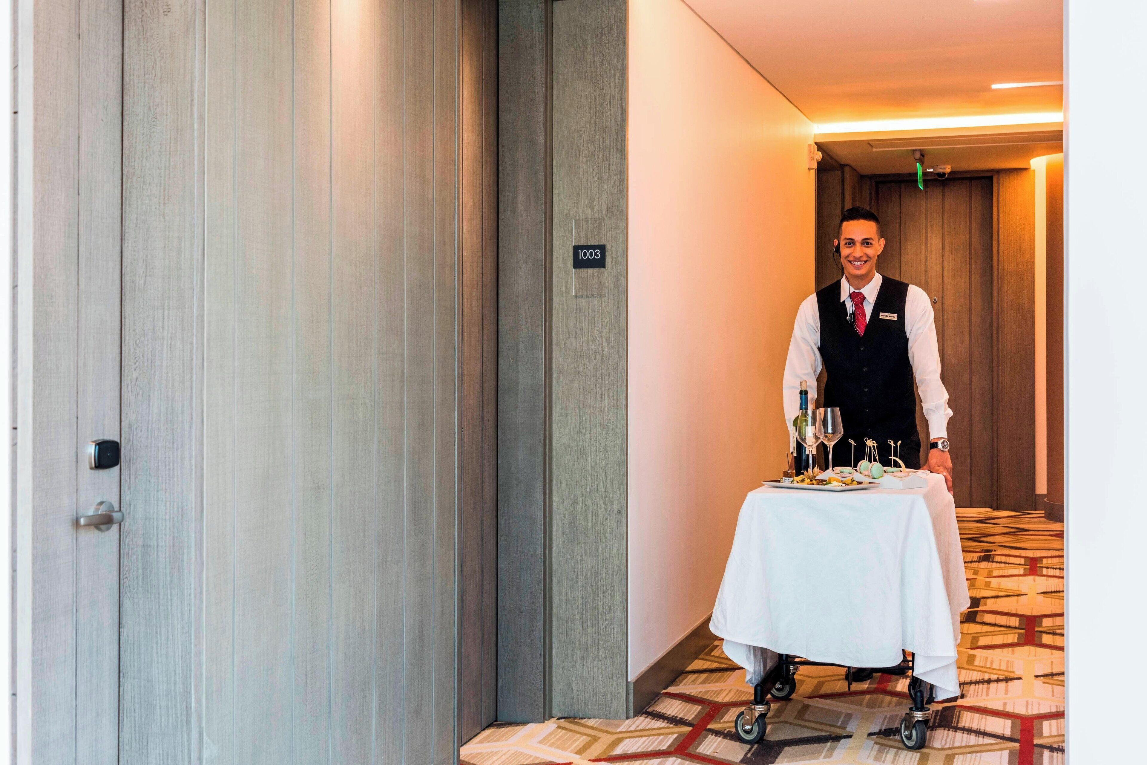 A hotel corridor with a wooden door marked "1003", a white table with a tray of food and drinks, and a carpeted floor.