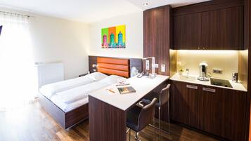 Une chambre d’hôtel comprenant un lit, un bureau, une chaise, un petit coin cuisine et une œuvre d’art colorée accrochée au mur.