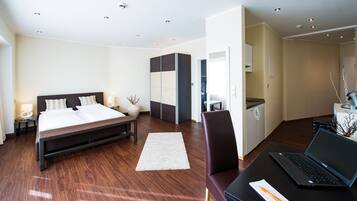 A modern hotel room with a large bed, a desk with a laptop, a chair, and a small kitchenette.