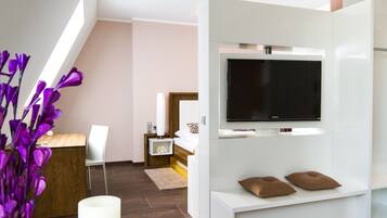 A modern hotel room with a wooden desk, a white chair, a bed with a zebra-patterned pillow, a TV mounted on a white wall, and a large window with natural light.