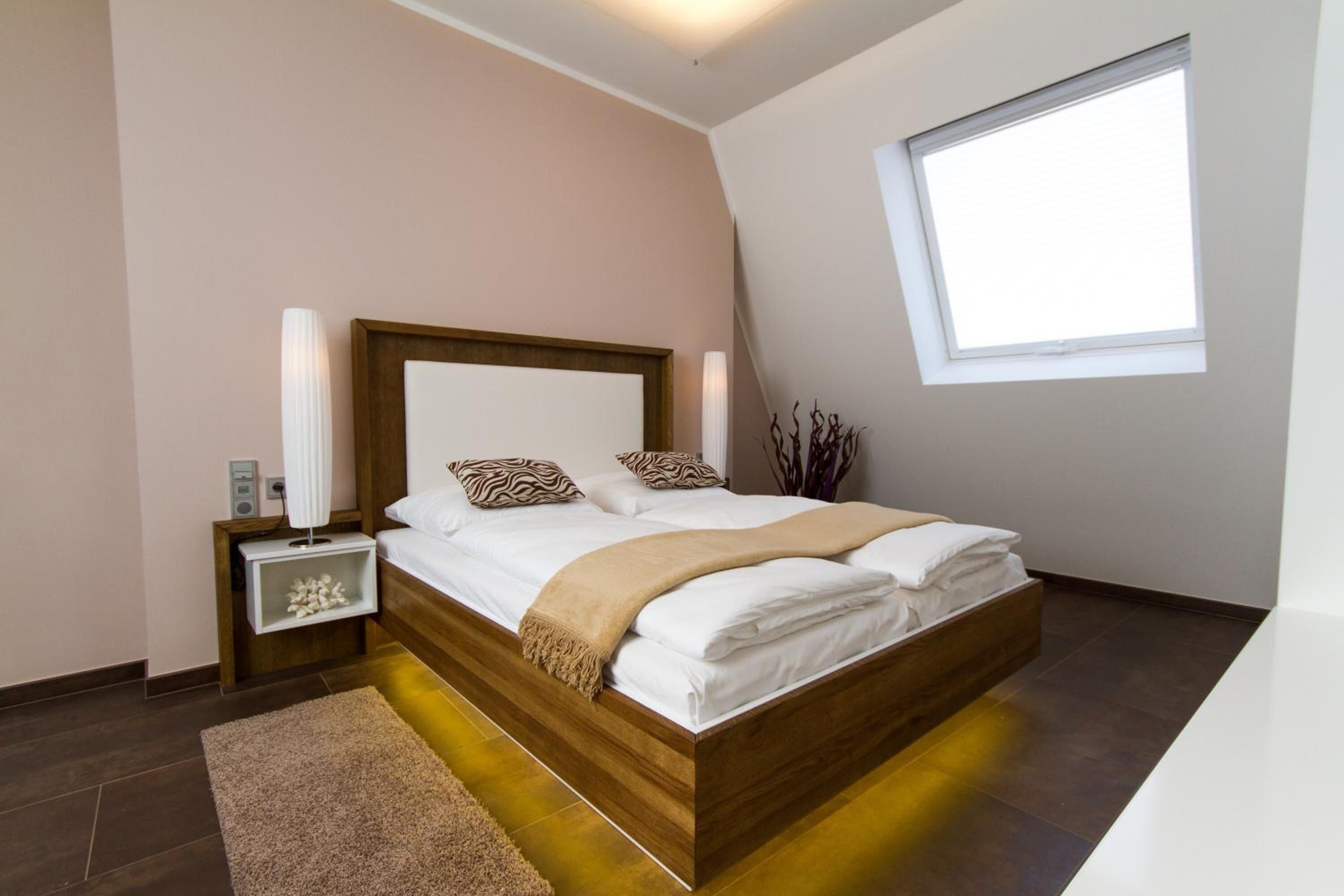 A modern bedroom with a wooden bed, white linens, a bedside table with a lamp, and a window with blinds.
