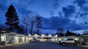 A motel with multiple rooms, a parking lot, and a large tree in the background. - Lakeview Lodge Motel (Lakeview)