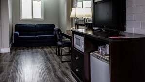 A hotel room with a dark wood floor, a blue sofa, a TV on a wooden cabinet, a lamp, and a window with blinds. - Lakeview Lodge Motel (Lakeview)