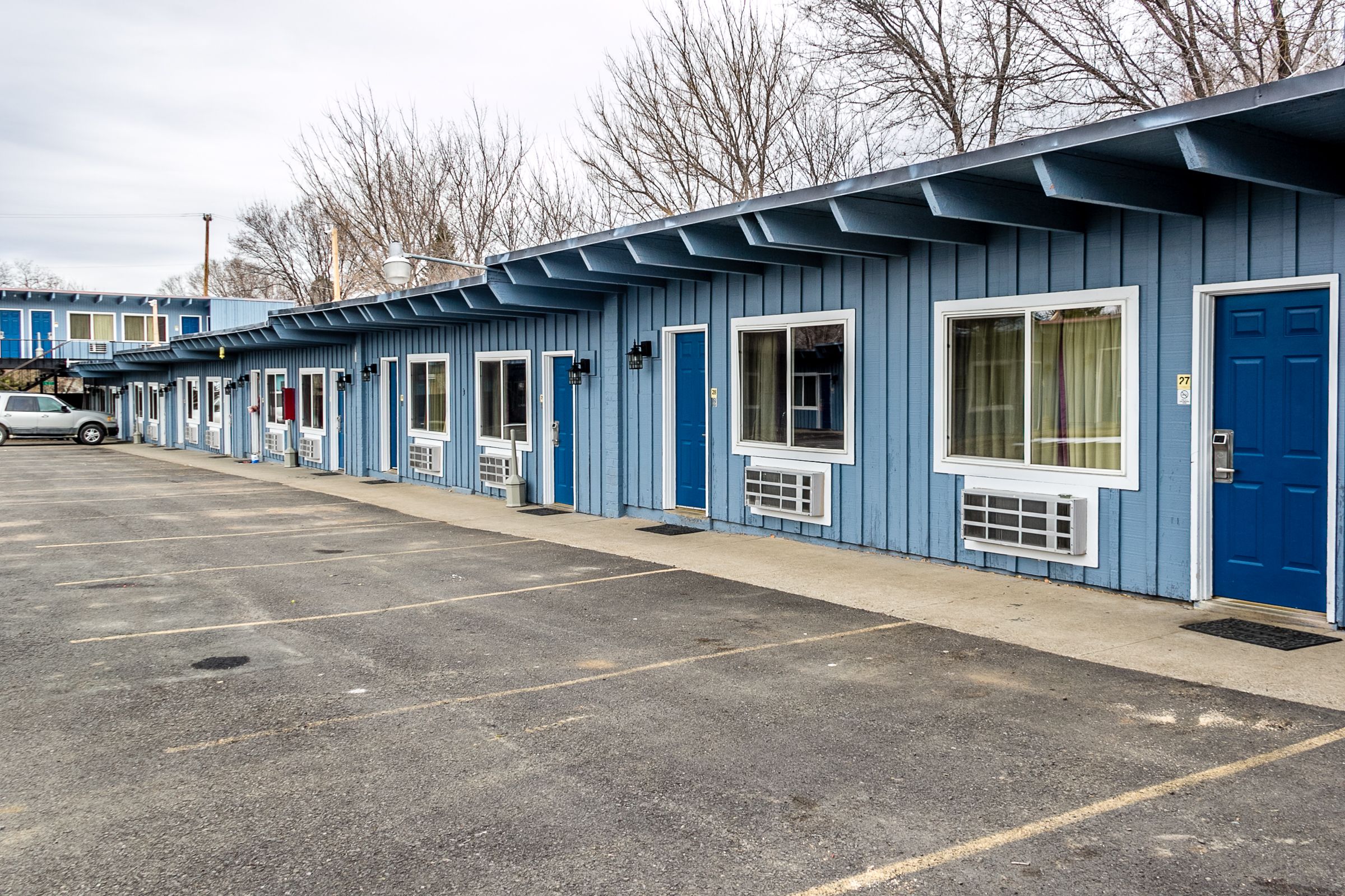 A row of blue motel rooms with individual windows and doors, a parking lot, and a car parked in front.