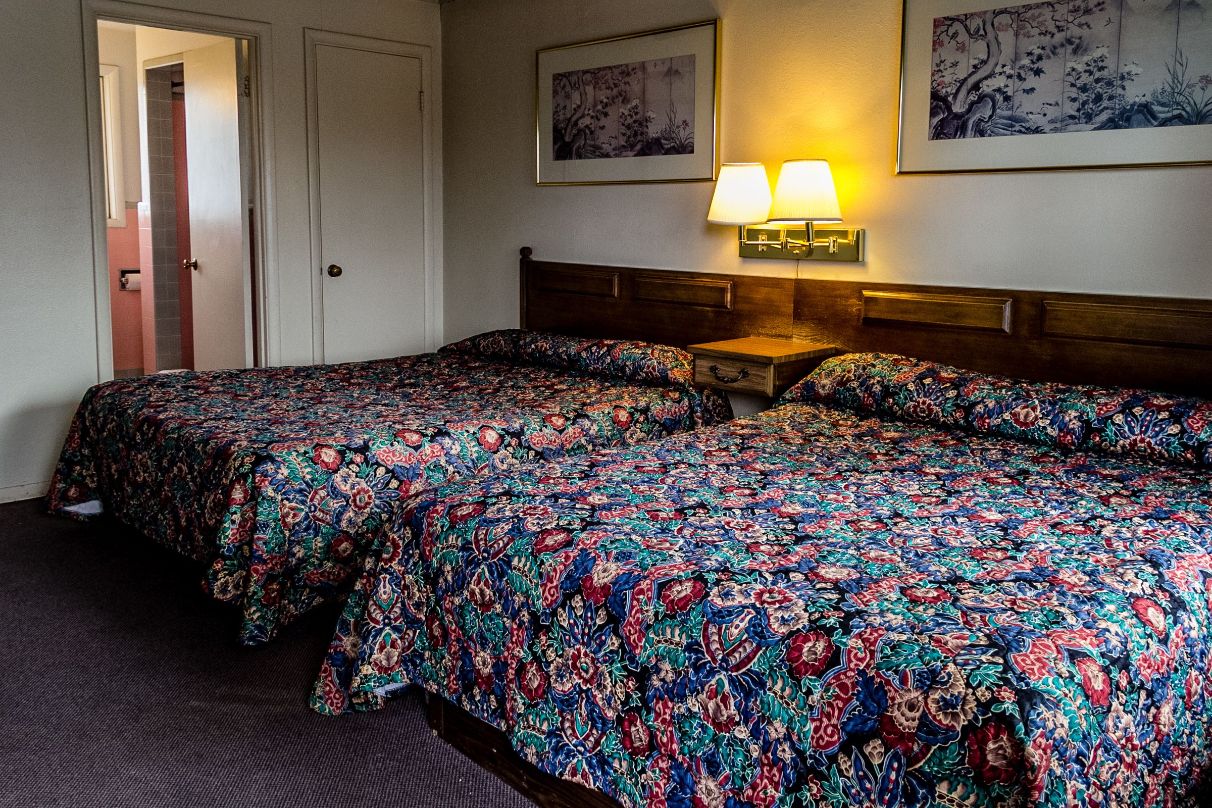 A hotel room with two beds, a nightstand with a lamp, and two framed artworks on the wall.