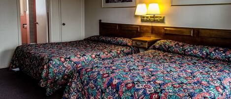 A hotel room with two beds, a nightstand with a lamp, and two framed artworks on the wall.