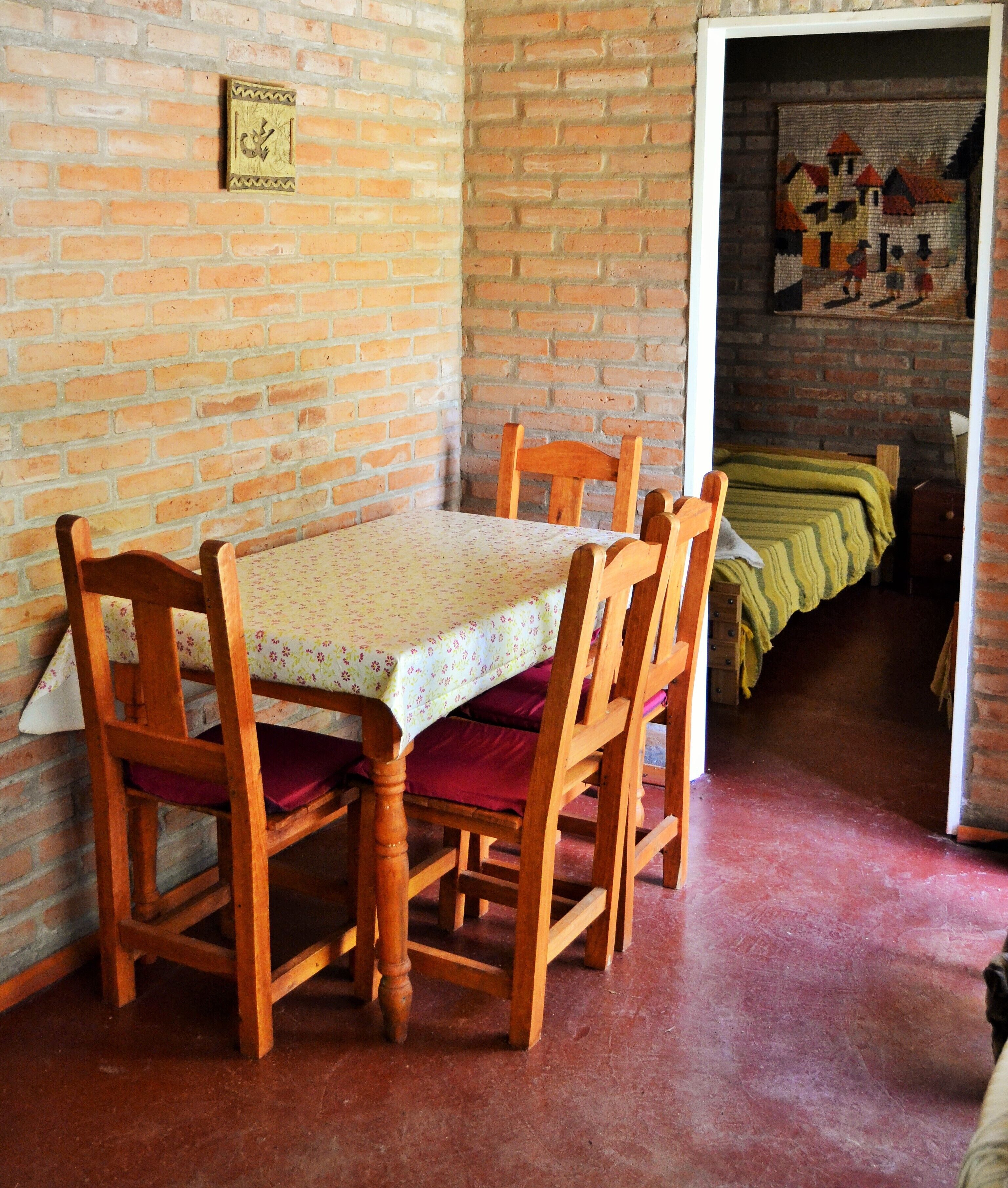 Un comedor con pared de ladrillo, una mesa con mantel floral, sillas de madera y suelo cubierto de alfombra roja.